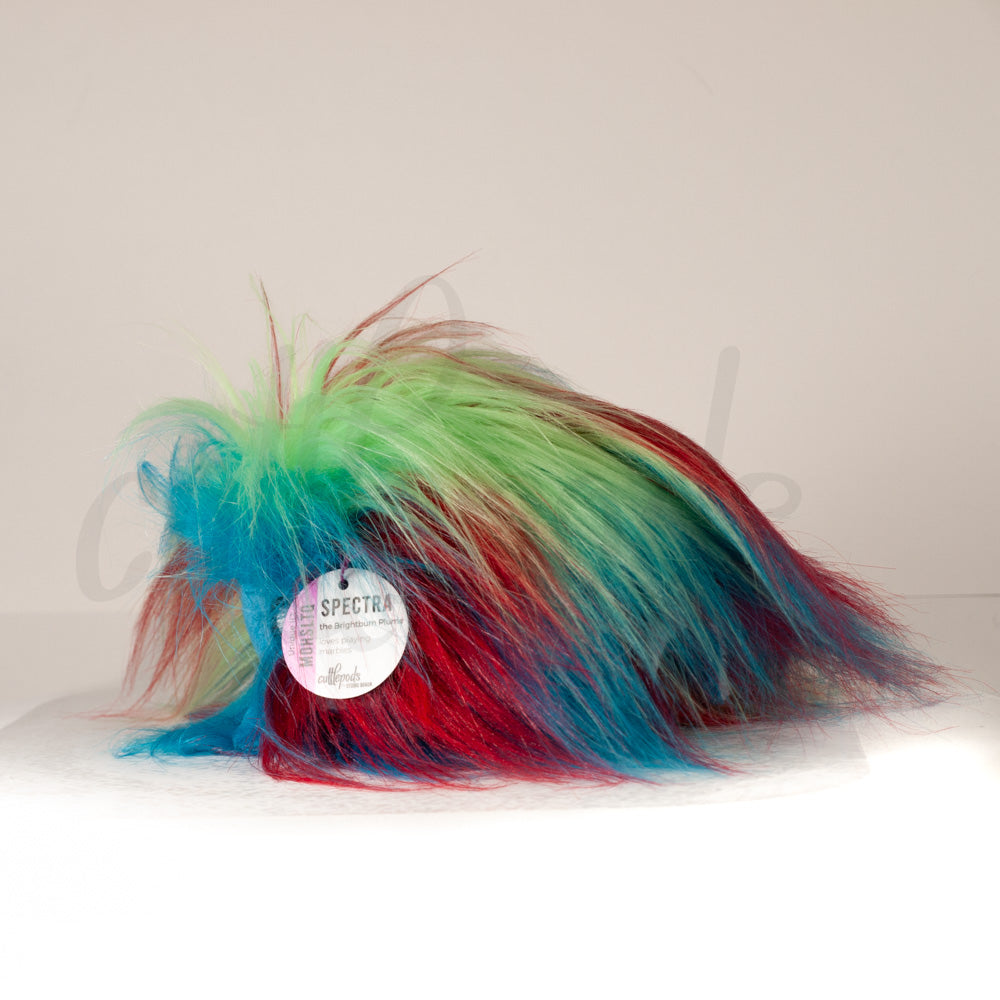 Slightly angled back view of a cuttlepod plush toy, highlighting two-tone ruffled fins, and a cuddly, furry body that comes to a soft point at the rear.