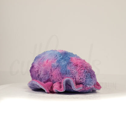 Slightly angled back view of a cuttlepod plush toy, highlighting two-tone ruffled fins, and a cuddly, furry body that comes to a soft point at the rear.