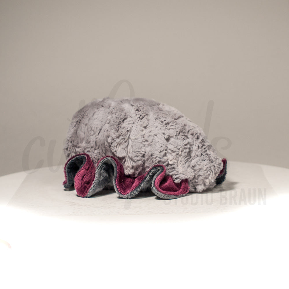 Slightly angled back view of a cuttlepod plush toy, highlighting two-tone ruffled fins, and a cuddly, furry body that comes to a soft point at the rear.