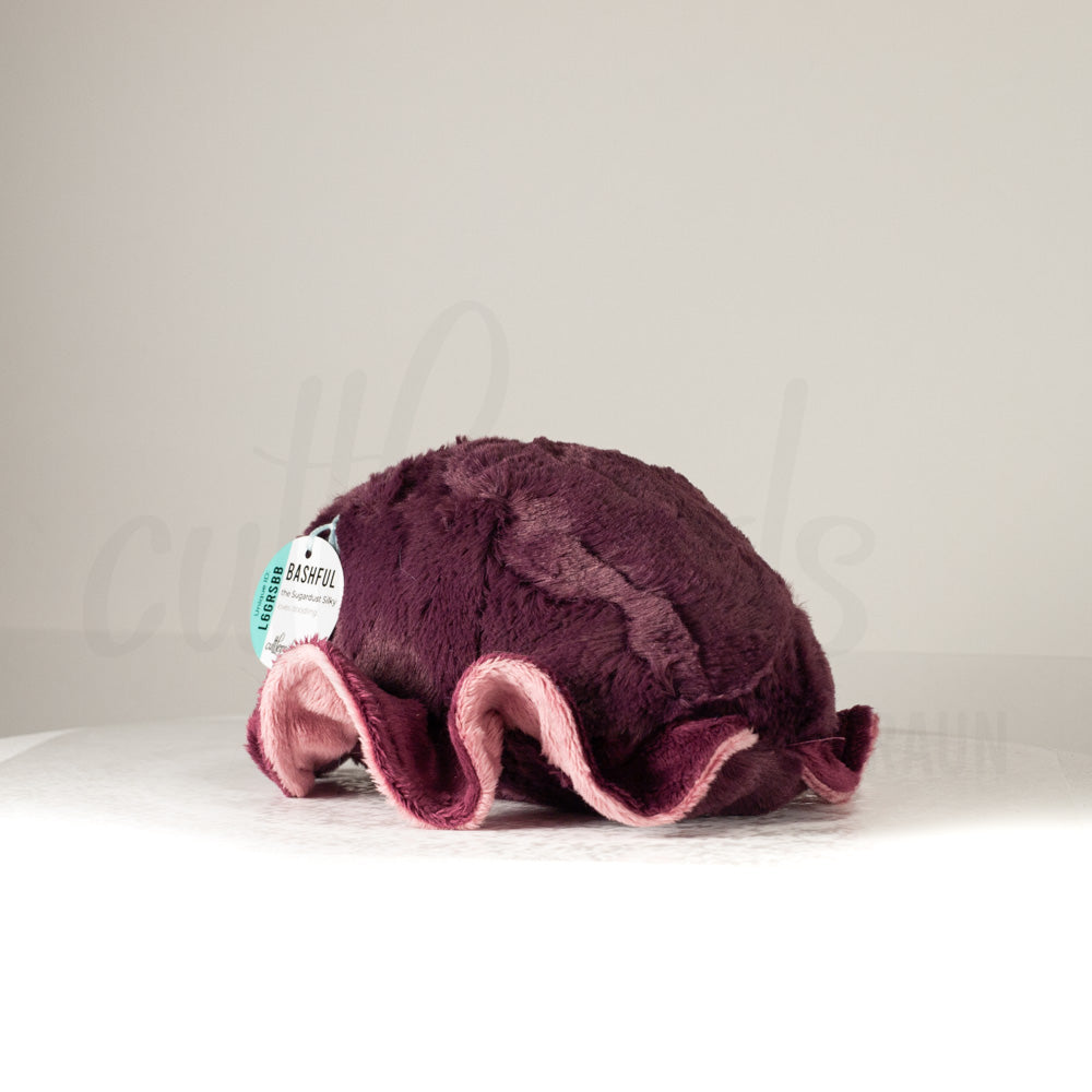 Slightly angled back view of a cuttlepod plush toy, highlighting two-tone ruffled fins, and a cuddly, furry body that comes to a soft point at the rear.