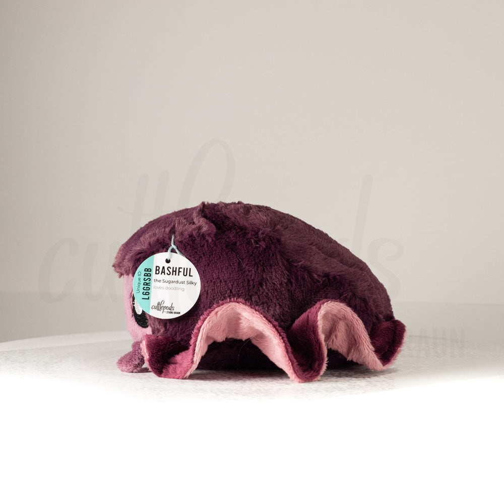 Side profile view of a cuttlepod plush toy, showcasing two-tone ruffled fins, and a cuddly, furry body.