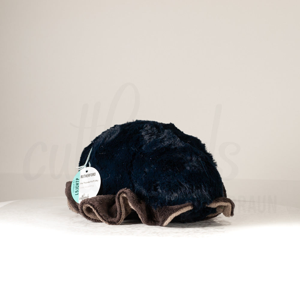 Slightly angled back view of a cuttlepod plush toy, highlighting two-tone ruffled fins, and a cuddly, furry body that comes to a soft point at the rear.