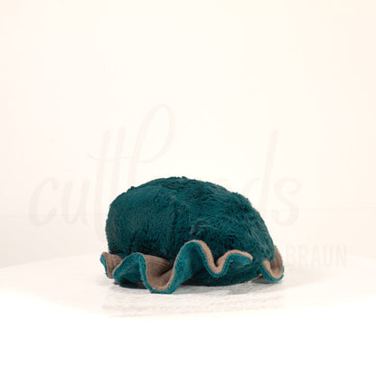 Slightly angled back view of a cuttlepod plush toy, highlighting two-tone ruffled fins, and a cuddly, furry body that comes to a soft point at the rear.